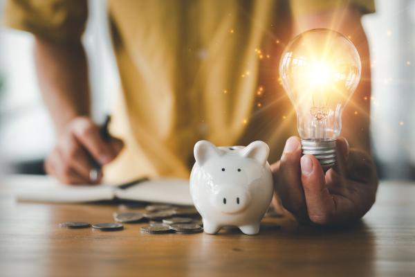 Persons hands holding a lit light bulb, next to a piggy bank