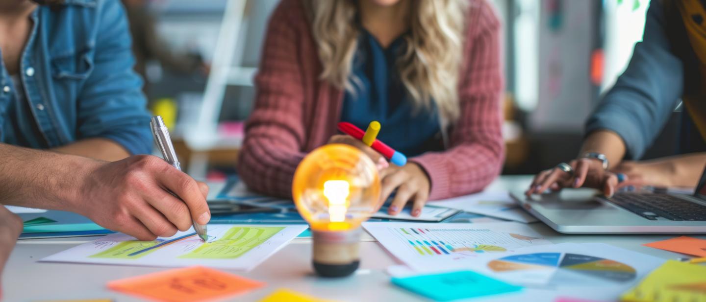 Group of 3 people sitting around a lightbulbs, looking at charts and graphs