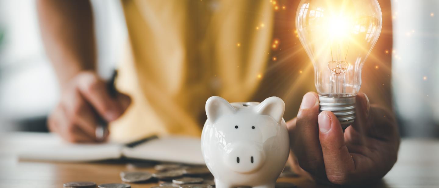 Persons hands holding a lit light bulb, next to a piggy bank
